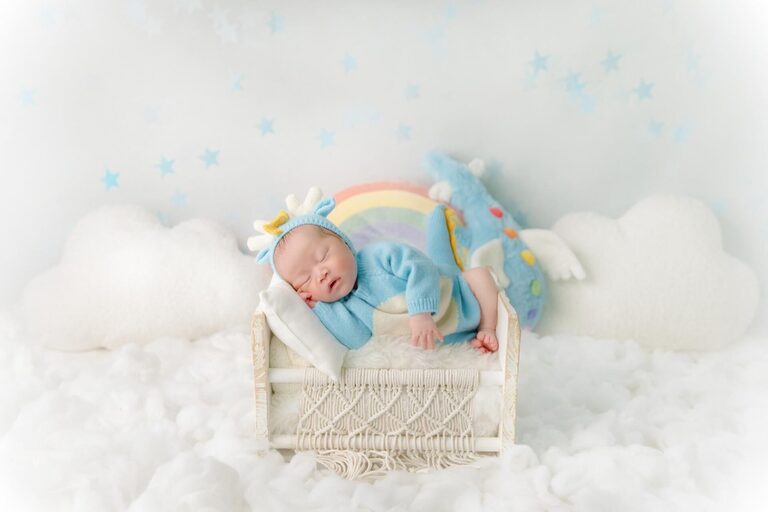 Newborn baby sleeping peacefully in a cozy setup with a soft pastel rainbow background, fluffy white clouds, and a whimsical blue dinosaur plush toy