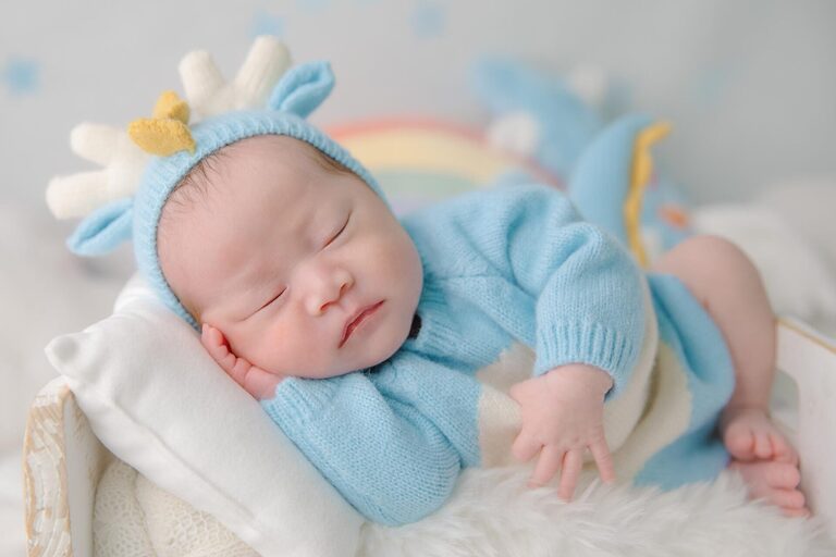 Newborn baby sleeping peacefully in a cozy setup with a soft pastel rainbow background, fluffy white clouds, and a whimsical blue dinosaur plush toy