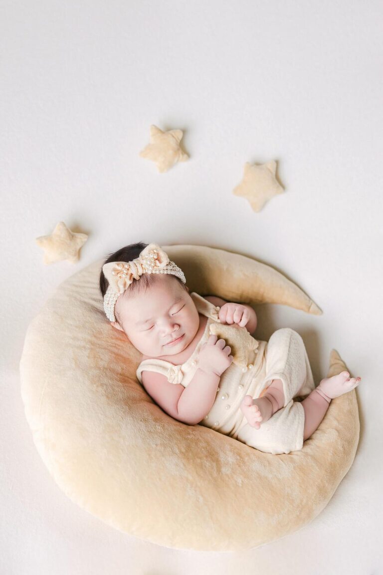 A newborn baby peacefully sleeps on a soft crescent moon-shaped cushion, surrounded by small star pillows, wearing a cream-colored outfit and a delicate pearl-embellished bow headband.