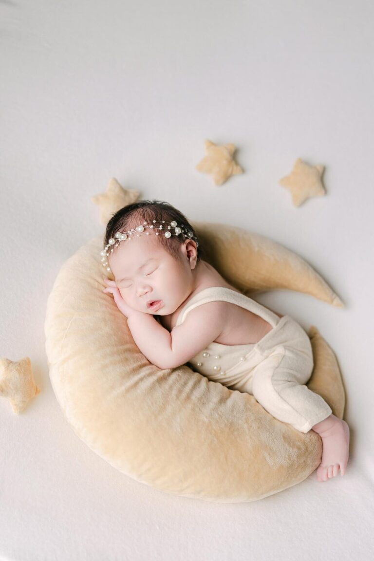 A newborn baby peacefully sleeps on a soft crescent moon-shaped cushion, surrounded by small star pillows, wearing a cream-colored outfit and a delicate pearl-embellished bow headband.
