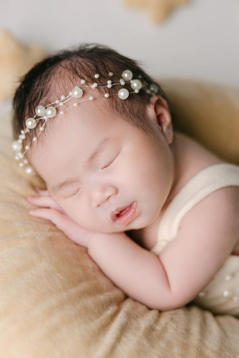 A newborn baby peacefully sleeps on a soft crescent moon-shaped cushion, surrounded by small star pillows, wearing a cream-colored outfit and a delicate pearl-embellished bow headband.