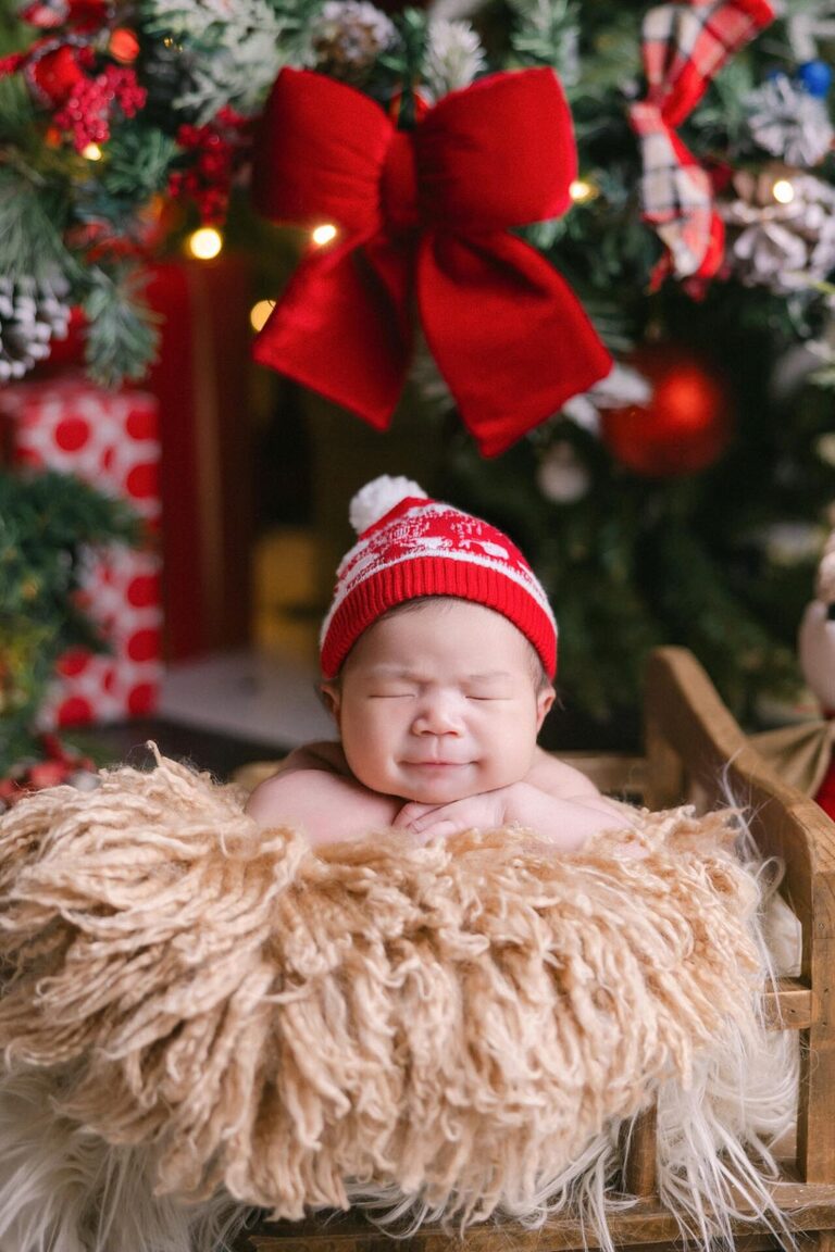 A newborn baby wearing a Santa hat sleeps peacefully in a cozy wooden sleigh with a fluffy blanket, surrounded by festive Christmas decorations, including a red bow, Christmas trees, and a reindeer plush