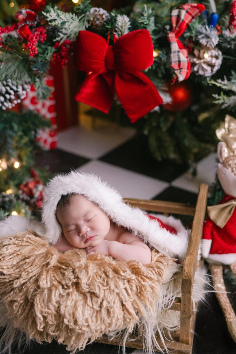 A newborn baby wearing a Santa hat sleeps peacefully in a cozy wooden sleigh with a fluffy blanket, surrounded by festive Christmas decorations, including a red bow, Christmas trees, and a reindeer plush.